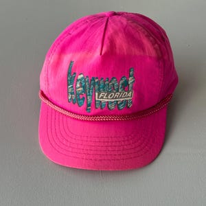 May include: A bright neon pink baseball cap featuring a braided cord trim across the base of the crown. The front displays embroidered text that reads Key West with the word FLORIDA printed in a white rectangular box underneath the lettering.