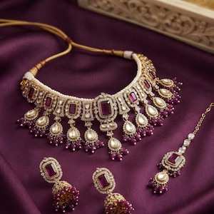 May include: A gold-toned jewelry set featuring a choker necklace, two bell-shaped earrings, and a maang tikka. The pieces are adorned with rectangular purple stones, clear crystals, and small white pearls, with dangling purple bead accents.