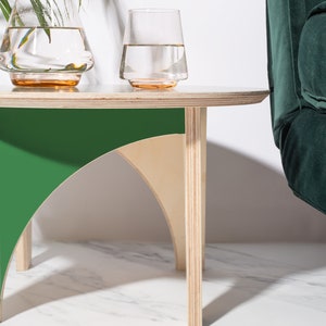 Może przedstawiać: Nowoczesny, minimalistyczny stolik boczny z zielonym i naturalnym drewnianym designem. Stół ma okrągły blat i łukowate nogi. Na stole stoją dwa przezroczyste szklanki z wodą i szklana karafka. W tle zielony aksamitny fotel.