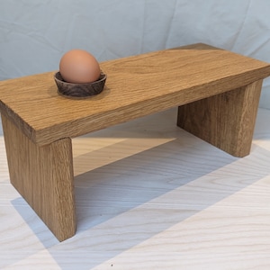 May include: A small, handcrafted wooden bench with a natural finish. An egg sits in a small, dark wooden bowl on top of the bench. The bench is made of light brown wood and has a simple, minimalist design.