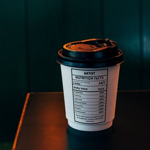 May include: A white to-go coffee cup with a black lid. The cup has a black and white label that reads "ARTIST NUTRITION FACTS" and lists the daily value of various qualities, such as originality, patience, ingenuity, and determination.