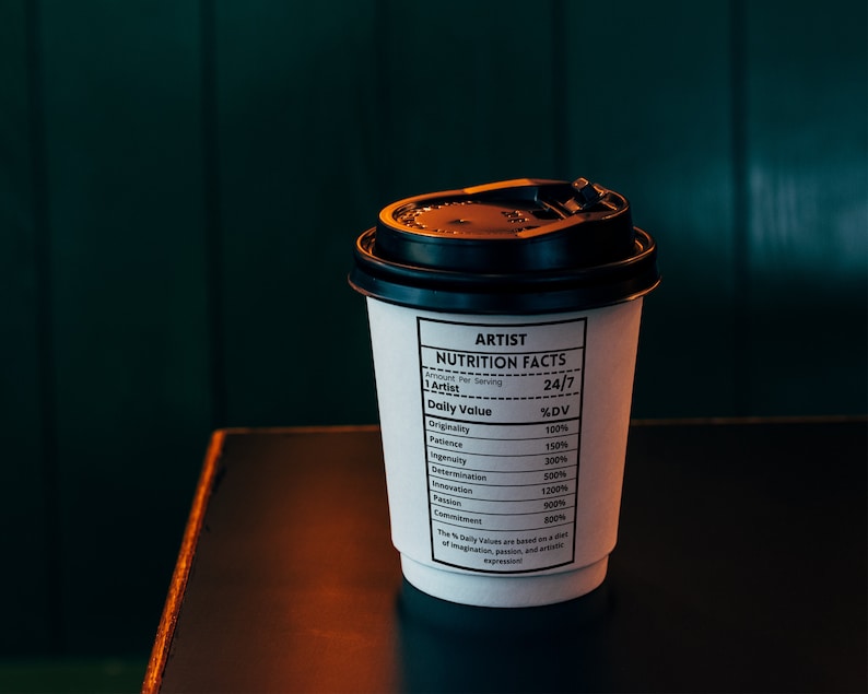 May include: A white to-go coffee cup with a black lid. The cup has a black and white label that reads "ARTIST NUTRITION FACTS" and lists the daily value of various qualities, such as originality, patience, ingenuity, and determination.