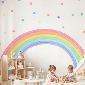 May include: A children's playroom with a large rainbow mural and colorful polka dots on the wall. Two young children are playing with wooden blocks on a large, round, white floor cushion. The room has light wood furniture.