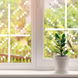 May include: A white window with a decorative design of white squares and lines. A small potted plant with green leaves sits on the windowsill. The background shows a blurred view of a green yard.