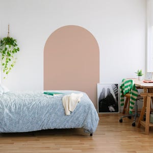May include: A bedroom interior featuring a bed with a blue patterned duvet, a wooden stool, and a desk with a green lamp. A large arch is painted on the wall in a neutral tone. A framed black and white photo is on the floor.
