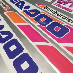 Może przedstawiać: Zestaw naklejek winylowych w kolorze białym, r&oacute;żowym, pomarańczowym i niebieskim z napisem "Sea-Doo" i "Bombardier".