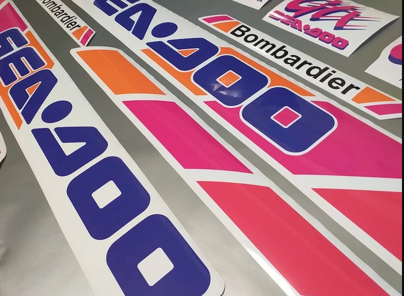 Może przedstawiać: Zestaw naklejek winylowych w kolorze białym, r&oacute;żowym, pomarańczowym i niebieskim z napisem "Sea-Doo" i "Bombardier".