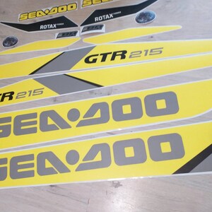 以下が含まれることがあります： 「Sea-Doo」と「GTR 215」の文字が入った、黄色と灰色のSea-Doo水上バイク用デカールセットです。