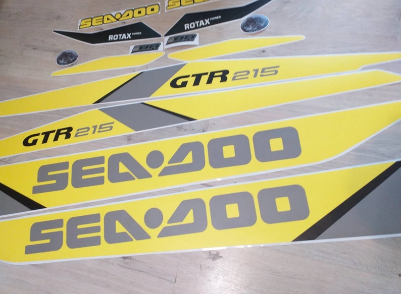 以下が含まれることがあります： 「Sea-Doo」と「GTR 215」の文字が入った、黄色と灰色のSea-Doo水上バイク用デカールセットです。
