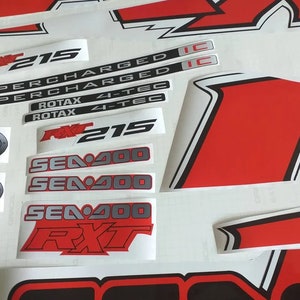 Peut inclure: Un ensemble d'autocollants noirs et blancs avec des accents rouges pour un bateau Sea-Doo. Les autocollants comprennent le texte "RXT 215", "SUPERCHARGED", "ROTAX 4-TEC" et "SEA-DOO".