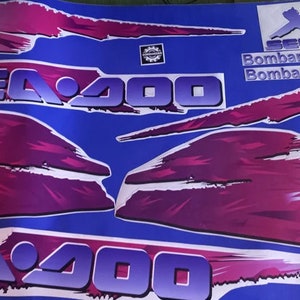Puede incluir: Un conjunto de calcomanías Sea-Doo XP Sea-Doo 200 de color rosa y morado con contornos blancos. Las calcomanías están sobre un fondo azul. El texto "Bombardier" aparece dos veces en las calcomanías.