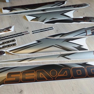 Puede incluir: Un conjunto de calcoman&iacute;as negras, blancas y plateadas para una moto acu&aacute;tica Sea-Doo. Las calcoman&iacute;as presentan el logotipo de Sea-Doo, el texto "Sea-Doo", "RX" y "Supercharged", y un logotipo de Rotax.