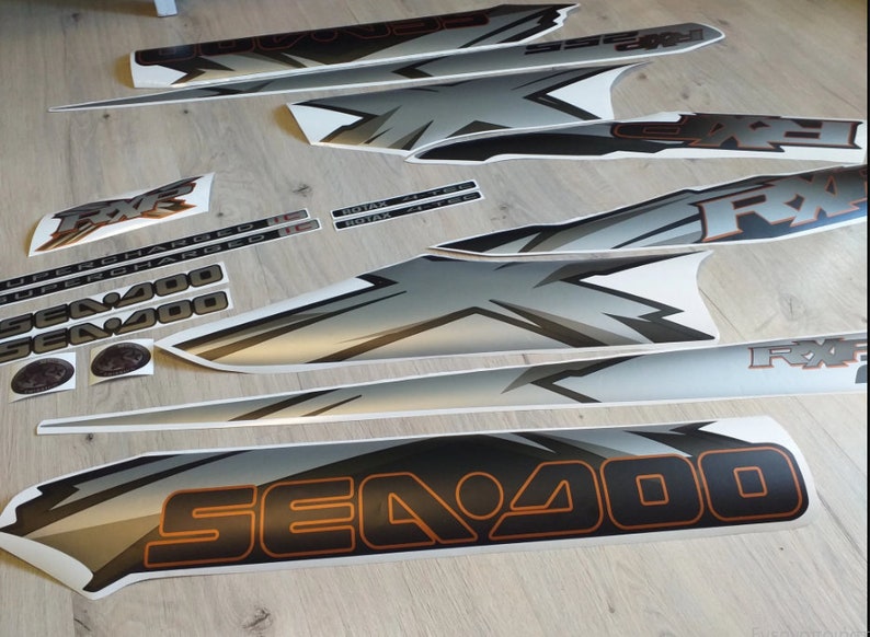 Puede incluir: Un conjunto de calcoman&iacute;as negras, blancas y plateadas para una moto acu&aacute;tica Sea-Doo. Las calcoman&iacute;as presentan el logotipo de Sea-Doo, el texto "Sea-Doo", "RX" y "Supercharged", y un logotipo de Rotax.