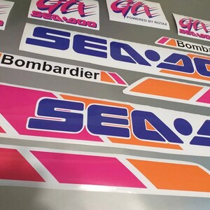 Może przedstawiać: Zestaw naklejek Sea-Doo w kolorze r&oacute;żowym, pomarańczowym i niebieskim. Naklejki przedstawiają logo Sea-Doo oraz tekst "Bombardier" i "Powered by Rotax".