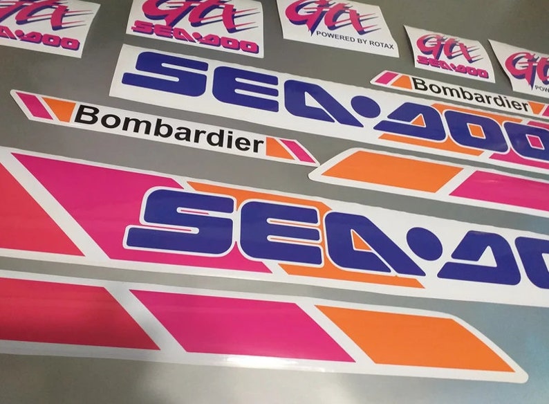Może przedstawiać: Zestaw naklejek Sea-Doo w kolorze r&oacute;żowym, pomarańczowym i niebieskim. Naklejki przedstawiają logo Sea-Doo oraz tekst "Bombardier" i "Powered by Rotax".