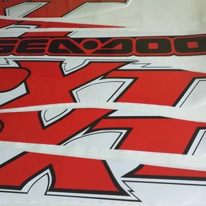 Peut inclure: Un ensemble d'autocollants en vinyle rouge et noir avec le mot "Sea-Doo" et un logo "RXT" stylis&eacute;. Les autocollants sont con&ccedil;us pour un engin nautique.