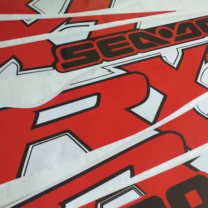 Peut inclure: Plusieurs feuilles d'autocollants en vinyle rouge et blanc avec les mots "Sea-Doo" et "RX" en lettres noires.