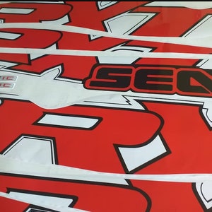 Peut inclure: Gros plan sur des autocollants graphiques rouges et blancs avec des contours noirs. Les autocollants pr&eacute;sentent les mots "Sea-Doo" et "R-X-R" sur un fond blanc. Les autocollants sont superpos&eacute;s.