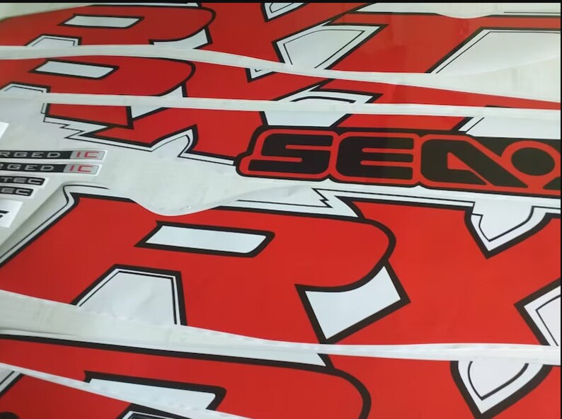 Peut inclure: Gros plan sur des autocollants graphiques rouges et blancs avec des contours noirs. Les autocollants pr&eacute;sentent les mots "Sea-Doo" et "R-X-R" sur un fond blanc. Les autocollants sont superpos&eacute;s.