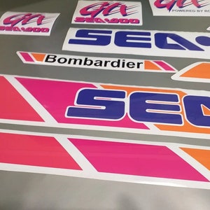 Może przedstawiać: Zestaw winylowych naklejek Sea-Doo do skuter&oacute;w wodnych w kolorze r&oacute;żowym, niebieskim, pomarańczowym i białym. Naklejki przedstawiają logo Sea-Doo i napis "Bombardier".