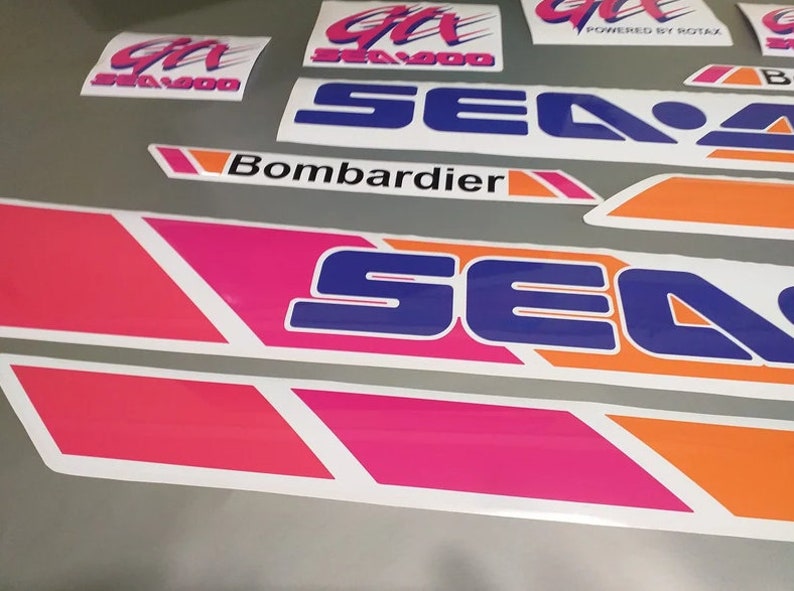 Może przedstawiać: Zestaw winylowych naklejek Sea-Doo do skuter&oacute;w wodnych w kolorze r&oacute;żowym, niebieskim, pomarańczowym i białym. Naklejki przedstawiają logo Sea-Doo i napis "Bombardier".