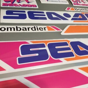 Może przedstawiać: Zestaw winylowych naklejek na motocykle wodne Sea-Doo z logo Sea-Doo w kolorze niebieskim i białym z pomarańczowymi i r&oacute;żowymi akcentami. Naklejki zawierają r&oacute;wnież tekst "Bombardier" i "Powered by Rotax".