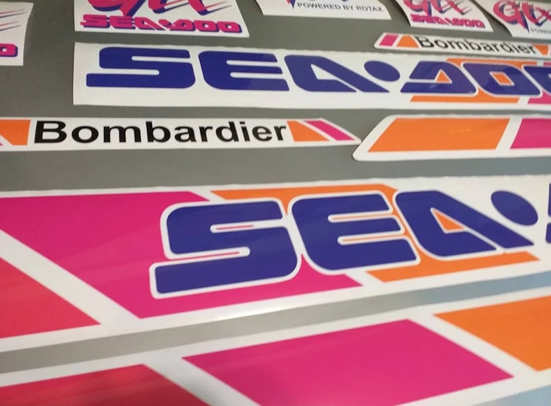 Może przedstawiać: Zestaw winylowych naklejek na motocykle wodne Sea-Doo z logo Sea-Doo w kolorze niebieskim i białym z pomarańczowymi i r&oacute;żowymi akcentami. Naklejki zawierają r&oacute;wnież tekst "Bombardier" i "Powered by Rotax".