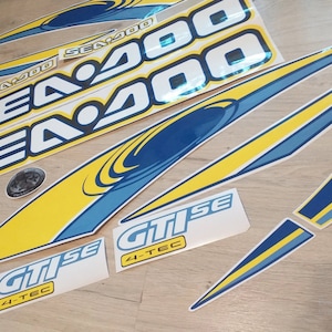 Peut inclure: Un ensemble d'autocollants Sea-Doo bleus et jaunes avec le texte "Sea-Doo" et "4-TEC GTI SE".