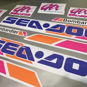 Może przedstawiać: Zestaw naklejek Sea-Doo w kolorze r&oacute;żowym, pomarańczowym i niebieskim. Naklejki przedstawiają logo Sea-Doo oraz tekst "Sea-Doo" i "Bombardier".