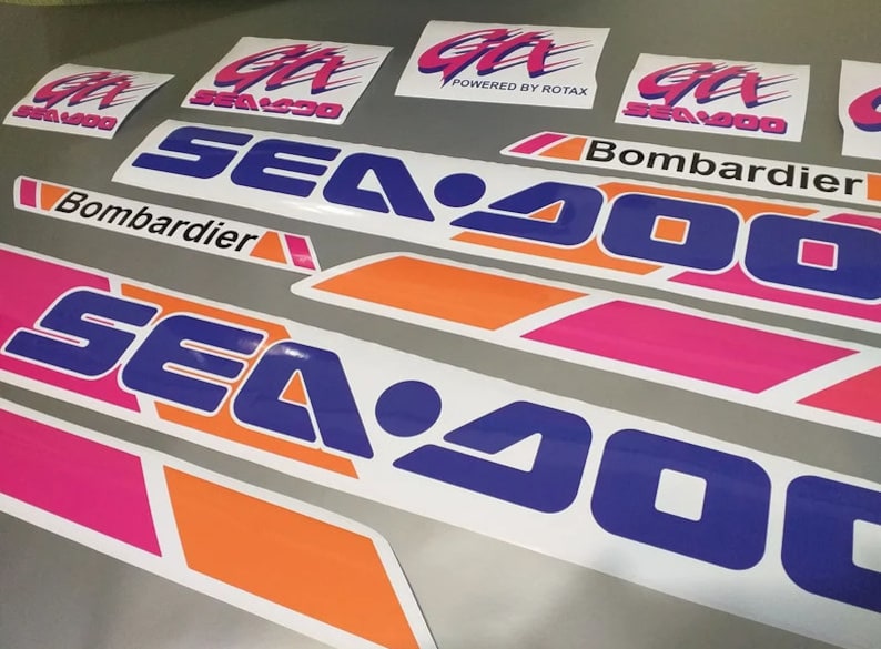 Może przedstawiać: Zestaw naklejek Sea-Doo w kolorze r&oacute;żowym, pomarańczowym i niebieskim. Naklejki przedstawiają logo Sea-Doo oraz tekst "Sea-Doo" i "Bombardier".
