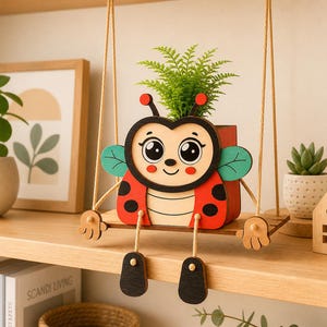 Puede incluir: Una maceta de mariquita de madera, suspendida por una cuerda. La mariquita tiene un cuerpo rojo con lunares negros, alas verdes y una cara sonriente. Contiene una pequeña planta verde, añadiendo un toque natural.