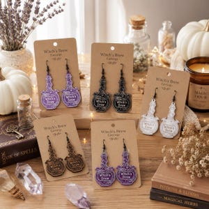 Pode incluir: Quatro pares de brincos pendentes em forma de frascos de poção, cada um num cartão castanho com a etiqueta Witch's Brew Earrings. Os brincos estão disponíveis em roxo, preto, branco e tons de madeira, com ganchos de metal preto.