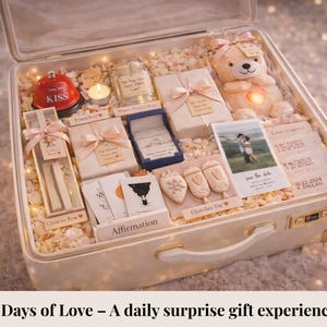 May include: A white suitcase filled with gifts, including a teddy bear, ring box, candles, and cards. The suitcase contains a variety of items, including a red bell that says "KISS". The text on the image says "7 Days of Love - A daily surprise gift experience".