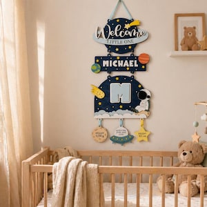 May include: A wooden nursery sign with a space theme. The sign says "Welcome Little One" and "MICHAEL". It includes a large letter "M", an astronaut, and planet and star decorations. The sign also includes the baby's weight, date, and height.