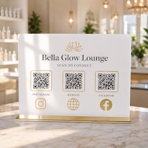 以下が含まれることがあります： 「Bella Glow Lounge」と「Scan to Connect」のテキストが表示された、金色のアクセントが付いた白いサイン。Instagram、Webサイト、FacebookのQRコードがあり、それぞれに金色のアイコンが付いています。サインは金色のベースにあります。