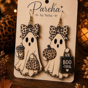 May include: A pair of wooden ghost earrings on a display card labeled Pareka by Neha. Each ghost features a leopard print bow, holding accessories with matching leopard print patterns. One ghost holds a coffee cup and purse, while the other holds a pumpkin and a black shopping bag with the text BOO-TIFUL and a heart icon.