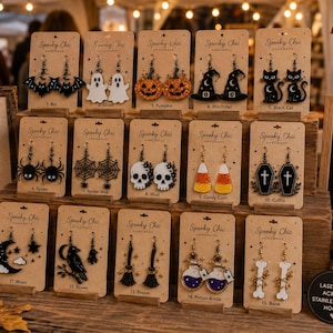 Op de afbeelding: Een display van vijftien paar lasergesneden acryloorbellen op merkkaartjes, met Halloween-motieven zoals vleermuizen, spoken, pompoenen, heksenhoeden, zwarte katten, spinnen, webben, schedels, snoepmaïs, doodskisten, manen, raven, bezems, toverdrankjes en botten.