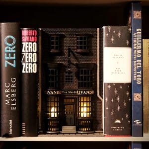 Puede incluir: Una maqueta en miniatura de una tienda con el texto "LIVANDE Fine Wands" en la fachada, ubicada entre libros en una estantería. La tienda tiene ventanas iluminadas y un exterior de ladrillo. Se ven libros con títulos como "ZERO" y "His Dark Materials".