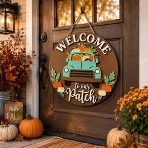 Op de afbeelding: Een rond houten deurbord met een blauwgroene vrachtwagen die oranje en witte pompoenen vervoert. Het bord toont de witte tekst WELCOME to our Patch. Het hangt aan een touw van jute tegen een donkerbruine deur met herfstdecoraties.