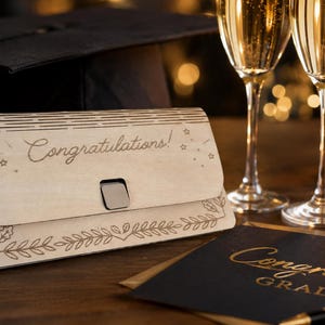 以下が含まれることがあります： 卒業祝いの木製カードホルダー。"Congratulations!"の文字が刻まれ、卒業帽子、シャンパンフルート、"Congrats Grad!"と書かれたカードが写っています。カードホルダーには葉の装飾が施されています。