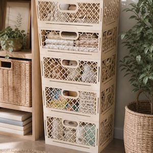 May include: A stack of five light-colored wooden storage crates with a decorative lattice design. Each crate has a handle and is filled with various items, including towels and toys. The crates are arranged against a wooden shelf and next to a potted plant.
