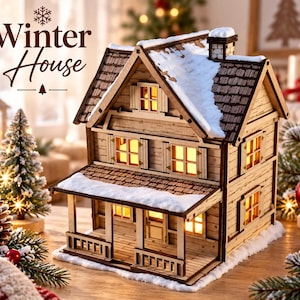 Puede incluir: Una casa de madera en miniatura de dos pisos con porche, ventanas iluminadas y detalles de nieve blanca en el techo y la base. El texto "Winter House" aparece en letra marrón oscura sobre la estructura, junto a un icono de copo.