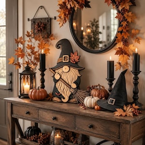 May include: A wooden gnome cutout wearing a black hat with a buckle and holding a broom, displayed alongside a black witch hat, orange and white pumpkins, black pillar candles in holders, a black lantern, and autumn maple leaf decorations.