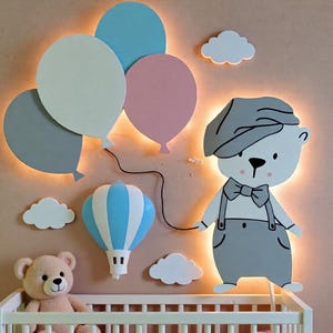 Puede incluir: Decoración de pared para guardería con un oso de dibujos animados con gorra, sosteniendo una cuerda de globo. Incluye globos, nubes y un globo aerostático en colores pastel. Un osito de peluche está sentado en una cuna blanca.