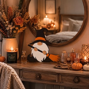 May include: A flat wooden gnome decoration featuring a black witch hat with an orange band, a white beard, and a broom with an orange brush. The gnome is positioned on a surface alongside a black candle labeled Pumpkin Spice and various glass bottles.