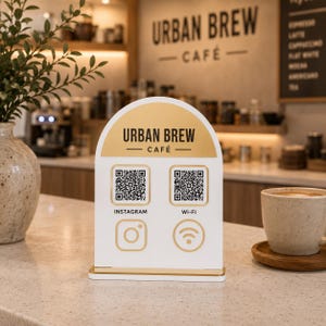 Op de afbeelding: Een wit en goudkleurig Urban Brew Cafe-bord met QR-codes voor Instagram en Wi-Fi. Het bord heeft een afgeronde bovenkant en een gouden basis. Rechts staat een kopje koffie op een houten onderzetter.