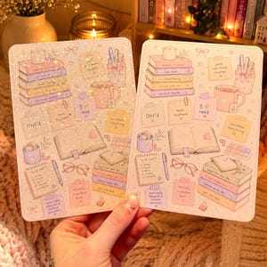 May include: Two rectangular sticker sheets featuring pastel illustrations of books, mugs, sticky notes, and reading accessories. The designs include text such as 'Just one more chapter', 'DNF'd', 'book boyfriend', and 'to be read' lists with various icons.