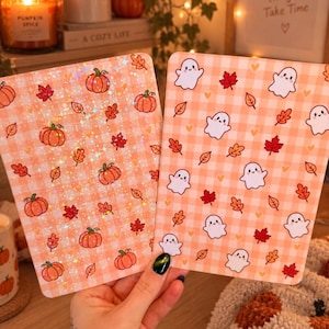 May include: Two rectangular cards with rounded corners featuring autumn themes. The left card has a holographic finish with orange pumpkins and red and orange leaves on a light orange gingham background. The right card shows white ghosts, red and orange leaves, and small yellow hearts on a light orange gingham background.