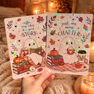 Può includere: Due stampe olografiche rettangolari con fantasmi dei cartoni animati. La stampa a sinistra dice coffee, cozy vibes, & good STORY con un fantasma che legge. La destra dice just one more CHAPTER con un fantasma, una tazza e libri impilati.