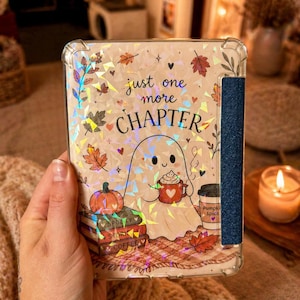May include: A clear e-reader case featuring a holographic design of a ghost holding a red mug, a pumpkin, a stack of books labelled Spooky, Story, and Coffee, and autumn leaves. The text reads just one more CHAPTER. A dark blue textured strap is attached.
